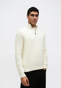 Λευκό πλεκτό πουλόβερ με φερμουάρ στον γιακά, με υφασμένα μανίκια και ριμπ τελείωμα. Φορεμένο με σκούρο παντελόνι, σε ένα απλό φόντο.