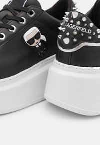 Černé kožené platformové tenisky mají zvýšenou bílou podrážku, stříbrné hroty na patě a detail v podobě kreslené nášivky po straně.