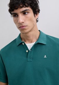 Polo de color verde oscuro con una tela texturizada, dos botones y un pequeño logo de calavera y huesos cruzados en el lado izquierdo del pecho.