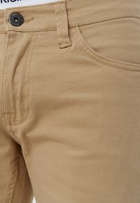 Beige bomuldsblandingsbukser med en glat tekstur, som har en frontlomme, metalknaplukning og syninger langs sømme.