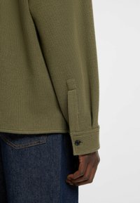 Olive green ribbed sweater with buttoned cuffs, featuring a textured fabric and a relaxed fit, paired with dark denim jeans.