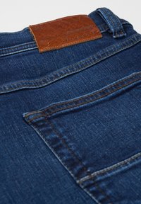 Close-up of dark blue denim jeans back pocket with brown leather patch labeled "bugatti" and belt loop visible.