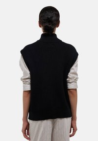 Gilet en maille côtelée noire avec col haut, porté par-dessus une chemise rayée à manches retroussées, présentant une silhouette décontractée.