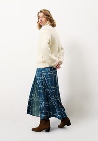 Crème fuzzy trui, lange blauwe rok met wit geometrisch patroon, en bruine haklaarzen, wat een gelaagde en textuurvolle outfit laat zien.