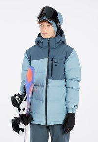 Blau-graue Pufferjacke mit Reißverschluss, Logo auf der Brust, kombiniert mit schwarzen Handschuhen, die ein buntes Snowboard mit weißen Bindungen halten.