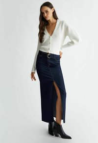 Cardigan bianco a coste con scollatura a V profonda, abbinato a una gonna in denim scuro con uno spacco frontale e texture in denim, indossato con stivali neri alla caviglia.