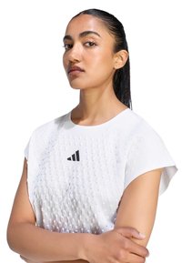 Jeune femme aux cheveux foncés attachés, portant un t-shirt blanc texturé Adidas, les bras croisés, regardant avec confiance sur le côté.
