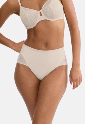 Kobieta w beżowym biustonoszu ozdobionym koronką i majtkach z wysokim stanem, stojąca z jedną ręką na biodrze na białym tle.