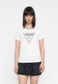 Woman with straight dark hair wears white Guess logo T-shirt and black denim mini skirt with large front pockets, standing against white background.