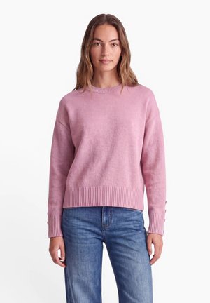 Kvinde iført en lyserød langærmet sweater med knapper ved manchetterne og blå jeans, stående mod en ensfarvet hvid baggrund.