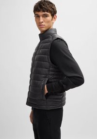 Black puffer vest, quilted design, sleeveless, with a high collar and side pockets. Worn over a long-sleeve black top. Smooth texture.