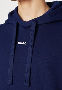 Námořnická modrá mikina vyrobená z měkké tkaniny, s kapucí na šňůrku a bílým logem "HUGO" na hrudi. Viditelné texturované šňůrky a švy.