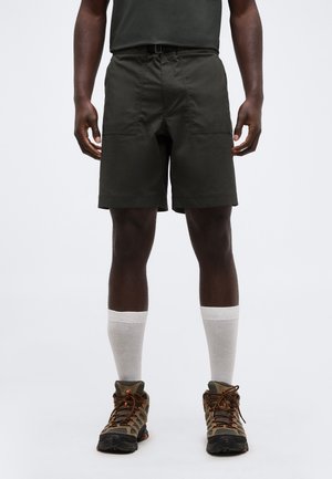 Άνδρας που φοράει σκούρα πράσινα σορτς, λευκές κάλτσες μέχρι τον αστράγαλο και καφέ παπούτσια πεζοπορίας, στέκεται μπροστά σε απλό φόντο.