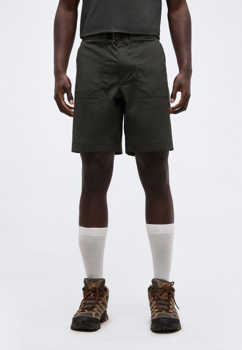 Άνδρας που φοράει σκούρα πράσινα σορτς, λευκές κάλτσες μέχρι τον αστράγαλο και καφέ παπούτσια πεζοπορίας, στέκεται μπροστά σε απλό φόντο.