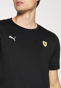 Černé bavlněné tričko s kulatým výstřihem. Na levé straně hrudi se nachází bílé logo Puma a emblém Ferrari se žlutými a červenými akcenty.
