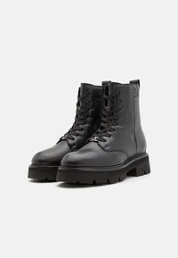 Bottes noires en cuir, mi-mollet, avec une semelle épaisse, un laçage à l'avant et des accents métalliques. Elles présentent une texture lisse et un talon renforcé.