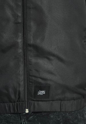 Must musträpiga musta jope, millel on esijalgsed tõmblukud ja elastne äär. Sisaldab väikest kangast silti alumises paremas nurgas.