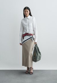 Biela košeľa na zapínanie s nazbieranou textúrou, béžová maxi sukňa so šikmým vzorom, červený šál zviazaný okolo pása, zelená taška, čierne lodičky.