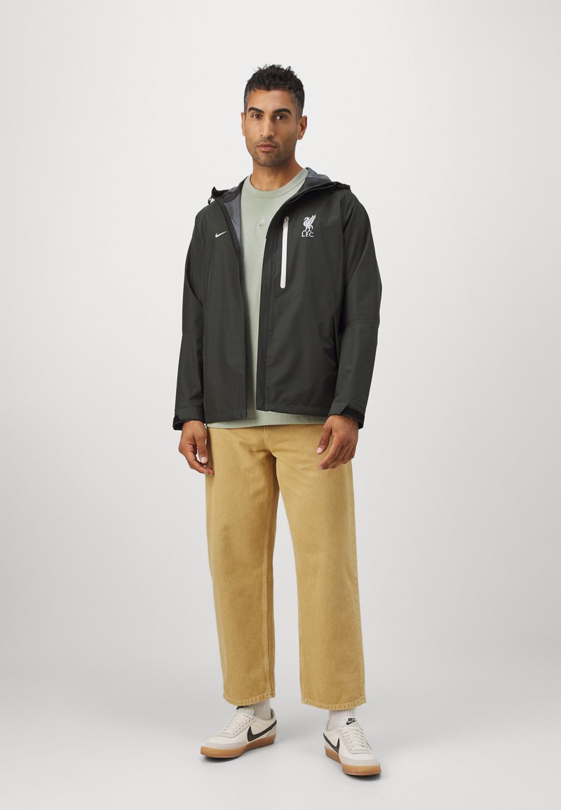 Schwarze, leichte Jacke mit grauen Akzenten und Logo, getragen über einem hellgrünen Hemd, kombiniert mit beigen Hosen und weißen Sneakers.