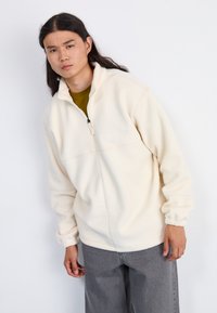 Kreemi fleece pullover, millel on veerandi tõmblukk, kõrge kaelus ja elastsed mansetid, koos hallide pükstega. Lihtne disain, ilma nähtavate mustriteta.