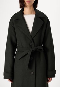 Μακρύ, σκούρο πράσινο μάλλινο παλτό με κουμπί μπροστά, oversized αθερίσκο και ζώνη που δένει στη μέση. Διαθέτει μεγάλα κουμπιά και φαρδιά μανσέτα.