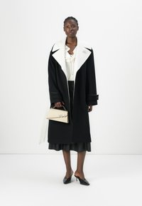 Manteau noir avec un col blanc, manches longues et une silouhette structurée. Le modèle tient un sac à main beige avec un logo. Chaussures noires et jupe.
