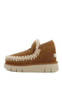 Bruine suede slipper met een beige geweven bies, gevoerde ronde neus en dikke lichtgekleurde zool. Beschikt over een logo op de hiel.