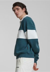 Sweatshirt colorat cu blocuri de culoare teal și alb, cu guler cu fermoar pe jumătate, materiale cu manșete și tiv ribbate. Țesătură moale cu o croială lejeră. Eticheta cu logo pe lateral.