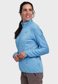 Helleblaue Zip-Jacke mit strukturiertem Design, seitlichen Taschen, hohem Kragen und einem dezenten Logo am Ärmel.