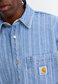 Lyseblå denimskjorte med lodrette hvide striber, trykknapper, en brystlomme og et gult Carhartt-logo-patch. Casual pasform og struktur.
