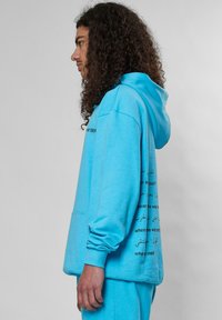 Sudadera turquesa de tejido suave, con un bolsillo frontal y un gran texto en negro en la manga izquierda y en la parte de atrás, con un corte relajado y capucha.