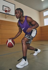 Lilla spordijalaga t-särk valge Nike logo ja hallid kamomustriga lühikesed püksid ning valged spordijalatsid. Korvpall hoitud paremas käes spordisaali põrandal.