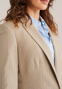 Smėlio spalvos dryžuotas švarkas iš tekstūruotos medžiagos, su plačiais atlapais ir subtiliomis mėlynomis linijomis, derinamas su šviesiai mėlyna marškinių apykakle.