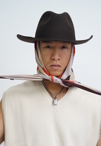 Black felt hat, beige sleeveless knit top with a v-neck, and a patterned silk scarf tied around the neck, featuring shades of cream, red, and brown.