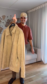 Beige ruskindsjakke med krave, udstyret med en sidelukning med lynlås. Modellen holder den på en bøjle og bærer en tekstureret rød skjorte og en beige kasket.