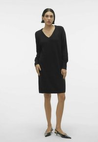 Abito nero a maglia con scollatura a V, maniche lunghe e vestibilità rilassata. Tessuto morbido, lunghezza al ginocchio, abbinato a sandali con tacco a punta.