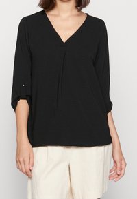 Sort bluse med V-udskæring og 3/4 ærmer. Blød, let tekstur med oprullede ærmer og en løs pasform. Båret med cremede shorts.