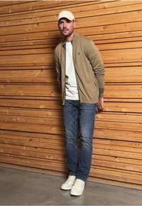 Jasno beżowa kurtka na zamek, noszona na białej koszuli, niebieskie dżinsy i białe trampki. Mężczyzna stoi na tle drewnianej paneli.