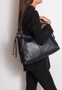 Marineblauwe leren handtas met een slouchy ontwerp, hardware in goudkleur, een ritsvak en een decoratieve sleutelhanger. Inclusief een verstelbare schouderriem.