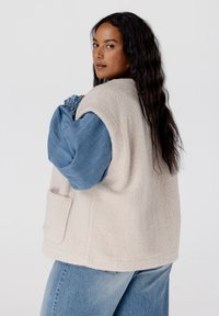 Crème fleece vest met een gestructureerde afwerking, voorzien van een voorzak, gedragen over een blauwe langarmtop. Het model heeft lang, golvend haar.