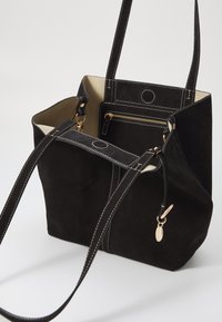 Geantă de tote din suede negru, cu cusături albe contrastante, având două mânere, un buzunar interior cu fermoar și un interior deschis la culoare.
