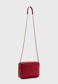 Bolso cruzado rectangular rojo con patrones geométricos texturizados y cadena dorada con almohadilla de cuero para el hombro, de pie contra un fondo blanco.