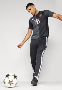 Svart träningströja med rosa detaljer, svarta träningsbyxor med vita sidostråk, och svarta fotbollsskor. En mönstrad fotboll ligger i närheten.