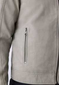 Gros plan d'une veste en suède gris clair avec une poche zippée verticale argentée sur le côté avant gauche et des détails de couture visibles.