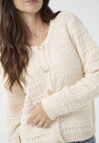 Cardigan tricotat de culoare crem deschis, cu un model texturat, având trei butoane decorative aurii și manșete cu nervuri.