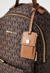 Brauner Rucksack mit Muster, ausgestattet mit einem goldenen Reißverschluss, Lederakzenten und einem Anhänger mit Logo. Das Design hat einen markanten Monogramm-Stil.