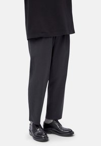 Parte inferiore del corpo di una persona che indossa pantaloni larghi neri, calzini grigi e scarpe eleganti in pelle nera su uno sfondo bianco.