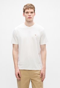 Camiseta branca de algodão com decote redondo, mangas curtas e pequeno logótipo vermelho no lado esquerdo do peito, combinada com calças bege.