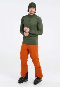 Olivenfarbener Langarmoberteil mit einem Viertel-Reißverschluss und flachen Nähten, kombiniert mit leuchtend orangefarbenen Cargo-Hosen und schwarzen Skischuhen.