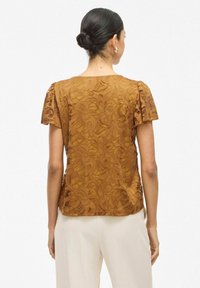 Ruda nėrinių bluzelė trumpomis rankovėmis, su gėlių raštais ir tekstūruotu paviršiumi. Spalvų detalė matoma ant pečių.